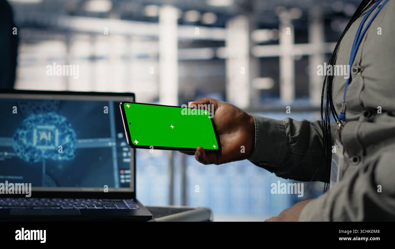 Data center worker uses green screen smartphone while deploying deep learning software. Server hub employee uses mockup mobile phone while training AI systems with datasets Stock Photo