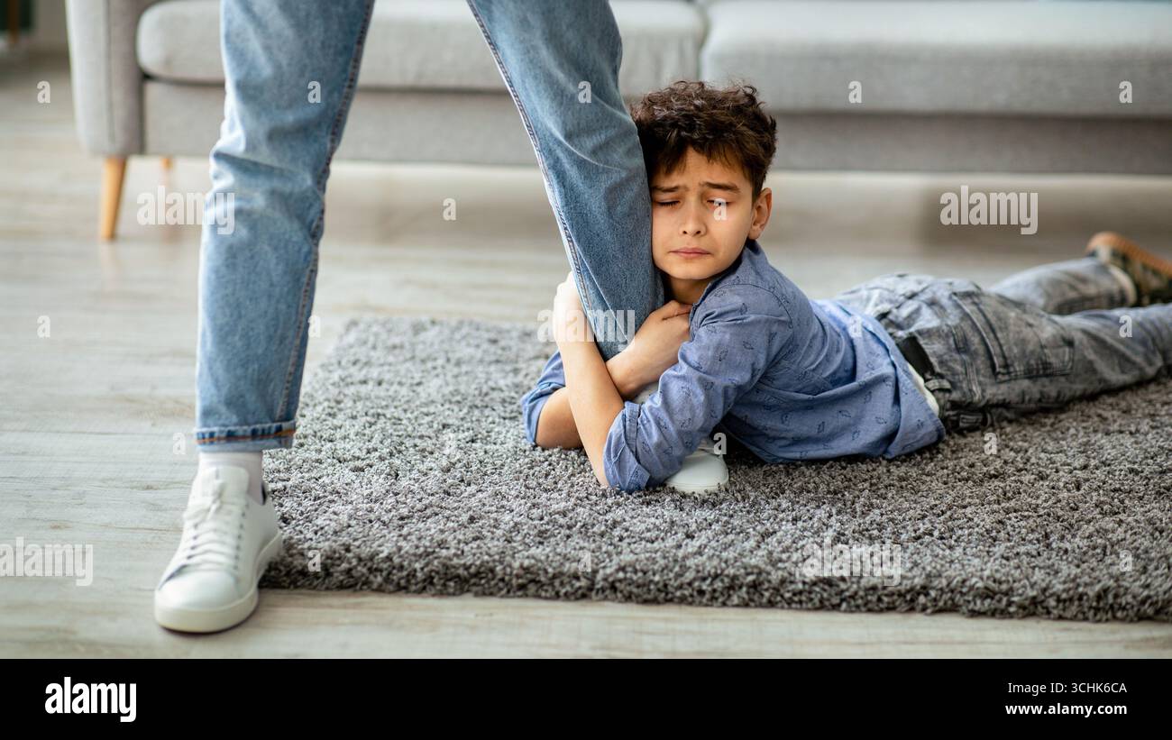 Don't go, dad. Upset boy embracing his father leg, begging daddy to stay with him, lying on the floor carpet Stock Photo