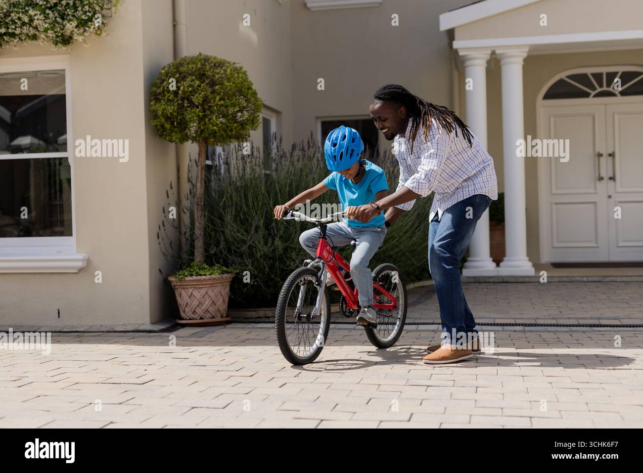 Father teaching son to ride bicycle in front of house, both smiling Stock Photo