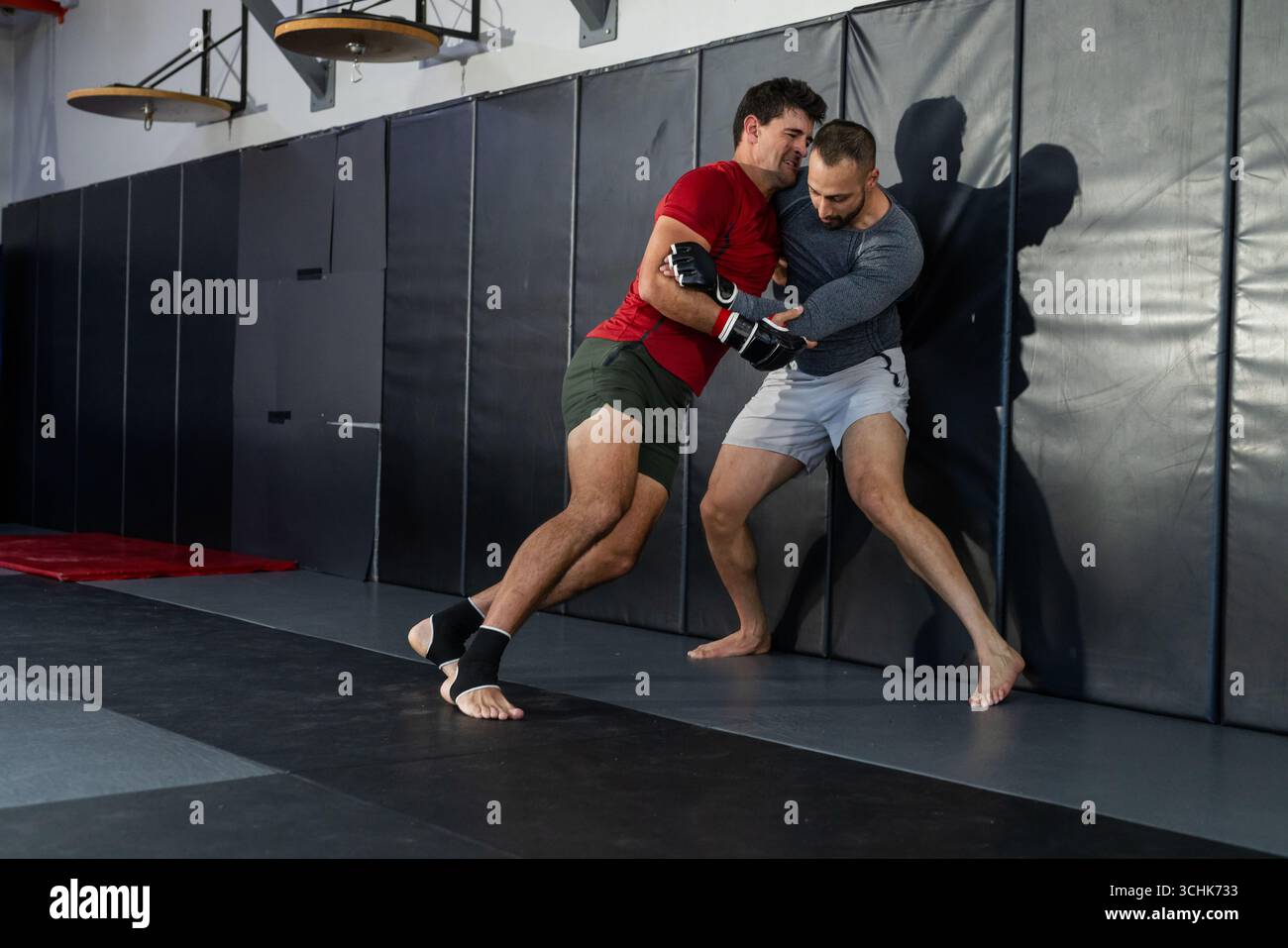 Diverse male training partners grappling on mats by padded wall at martial arts gym in gloves Stock Photo