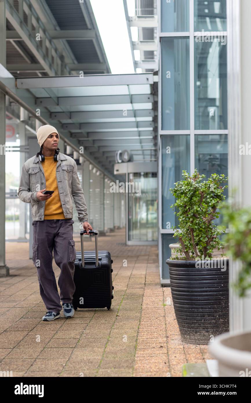 African american man pulling suitcase while using smartphone on walkway near planters Stock Photo