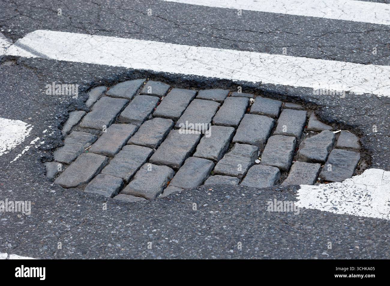 A New York City crosswalk pothole with a patch of missing asphalt reveals a cobblestone street underneath. Stock Photo