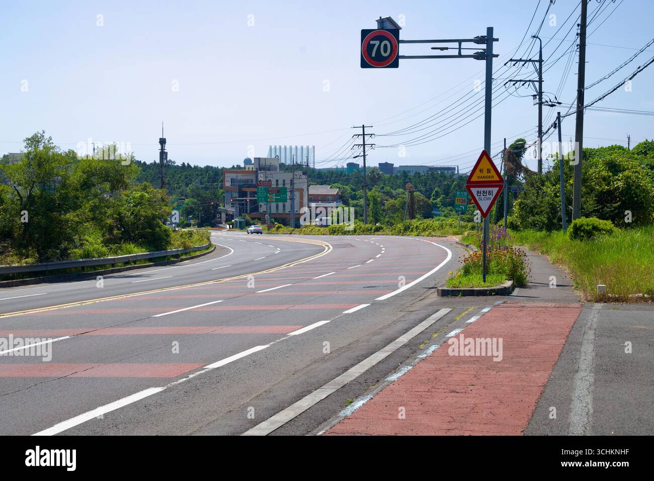 A 70‑kilometer speed sign and hazard warning stand by a sweeping curve on Iljuseo Road, with guardrails, utility poles, and distant buildings beyond t Stock Photo