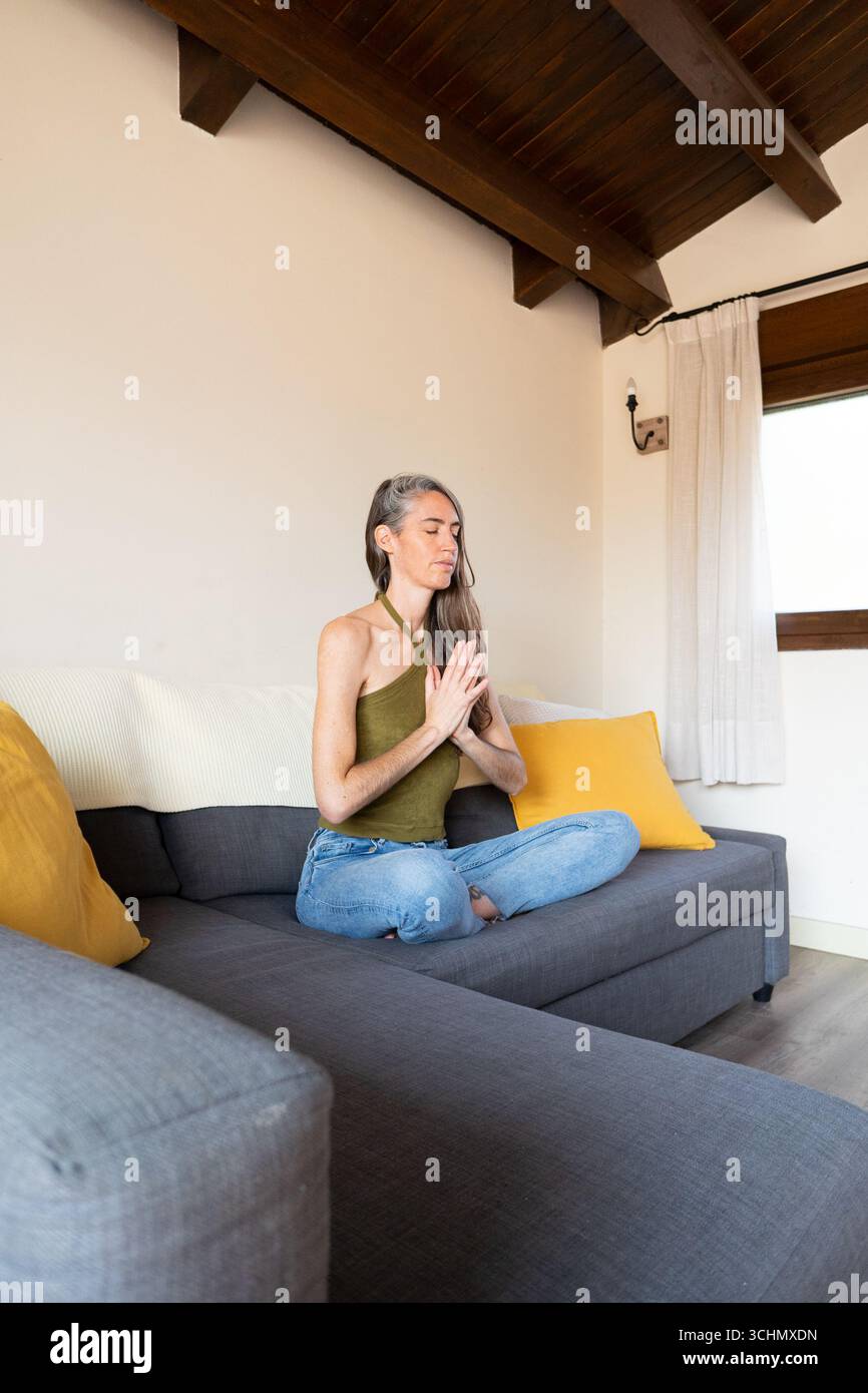Serene mature woman practicing meditation on comfortable sofa, embracing mindfulness and finding inner peace in the tranquility of her home Stock Photo
