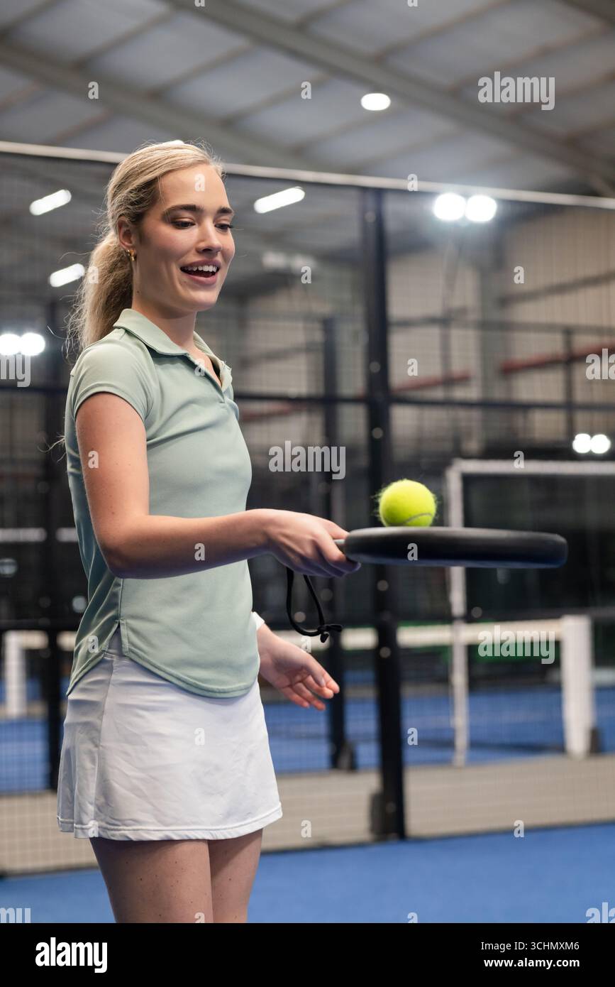 Female athlete holding padel racket balancing tennis ball on padel court near metal fencing, net Stock Photo
