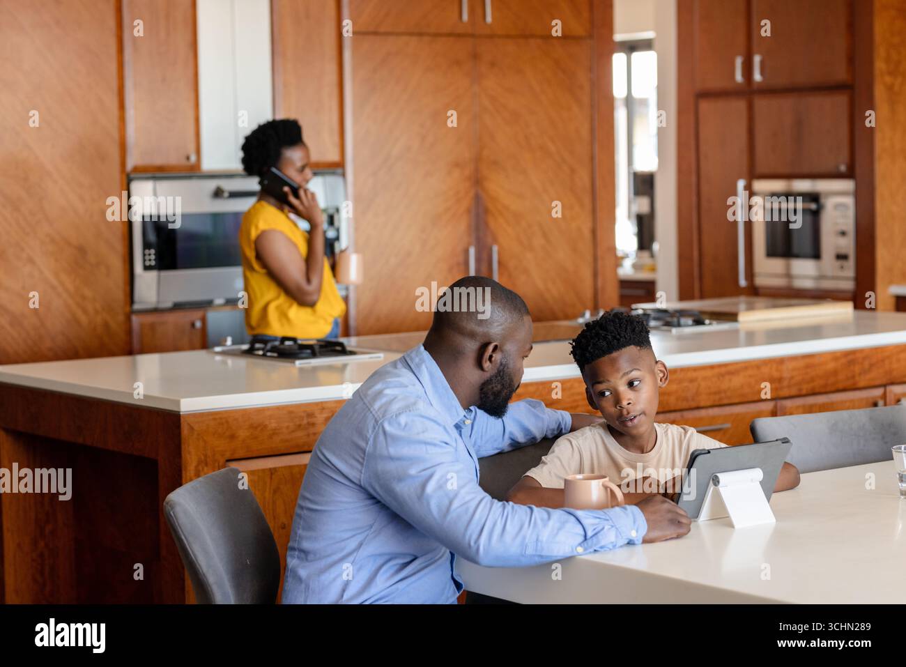 Father and son using tablet at kitchen table while mother talks on phone Stock Photo