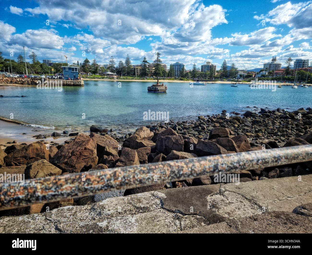 A scenic view of a rocky shoreline and calm bay with anchored boats, framed by a vibrant blue sky and city skyline in coastal Australia. - Smartphone Captured Stock Image