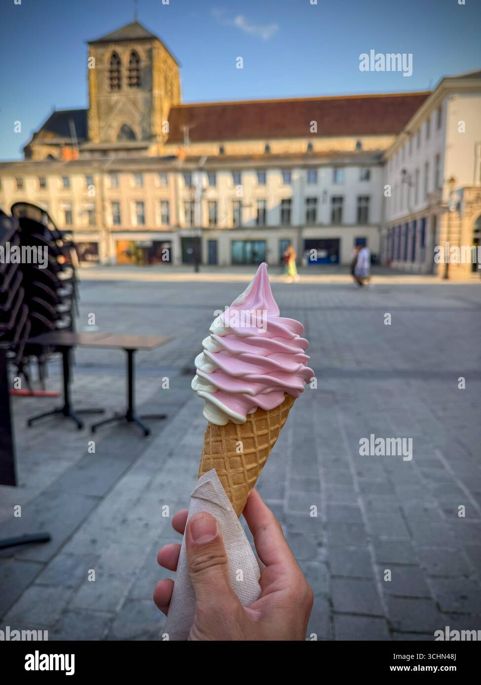 Hand holding a swirl ice cream cone in a town square, with historic buildings and architecural details in the background - Smartphone Captured Stock Image