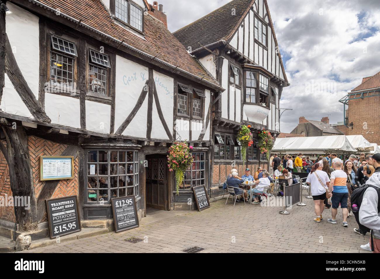 Gert & Henry’s Restaurant in York, England. A timber framed Tudor building with hanging baskets, outdoor diners, and traditional roast menu signage. Stock Photo