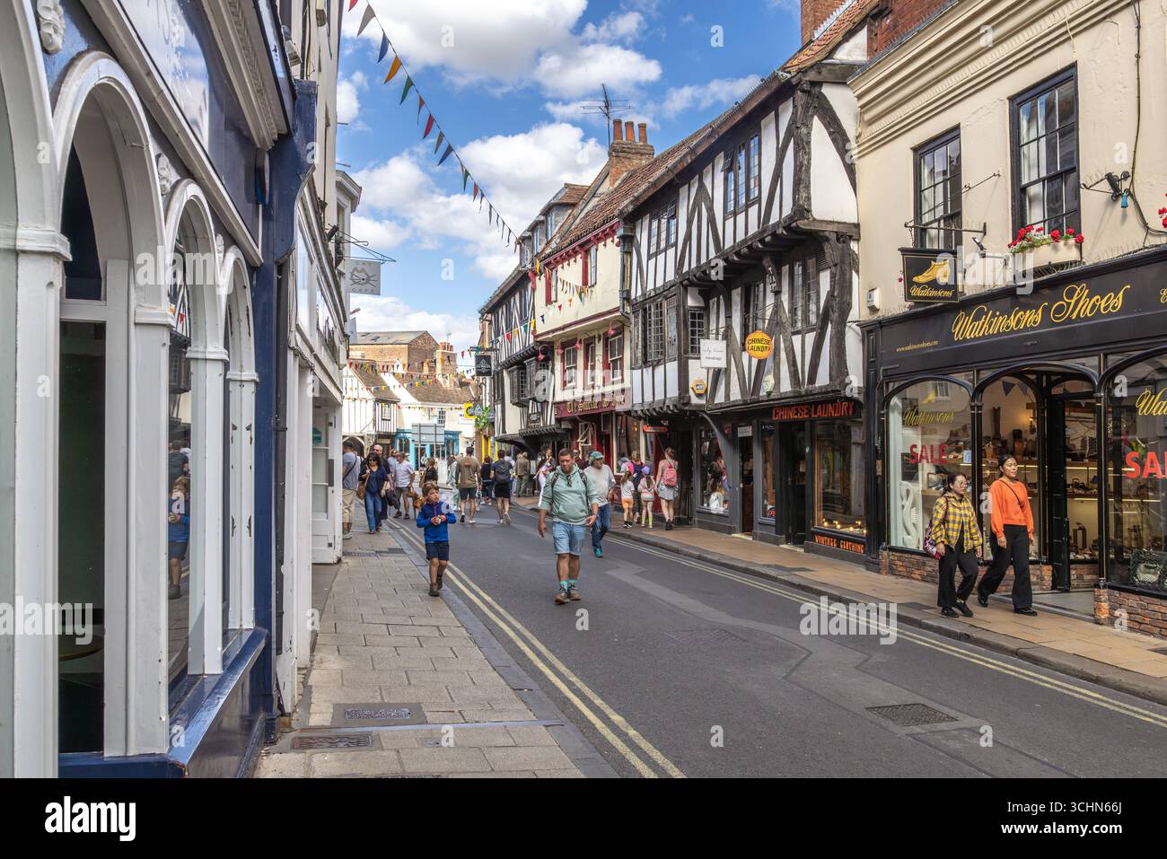 Pedestrians walk past timber-framed buildings and shops on a lively street in York, England, decorated with bunting and active with tourism. Stock Photo