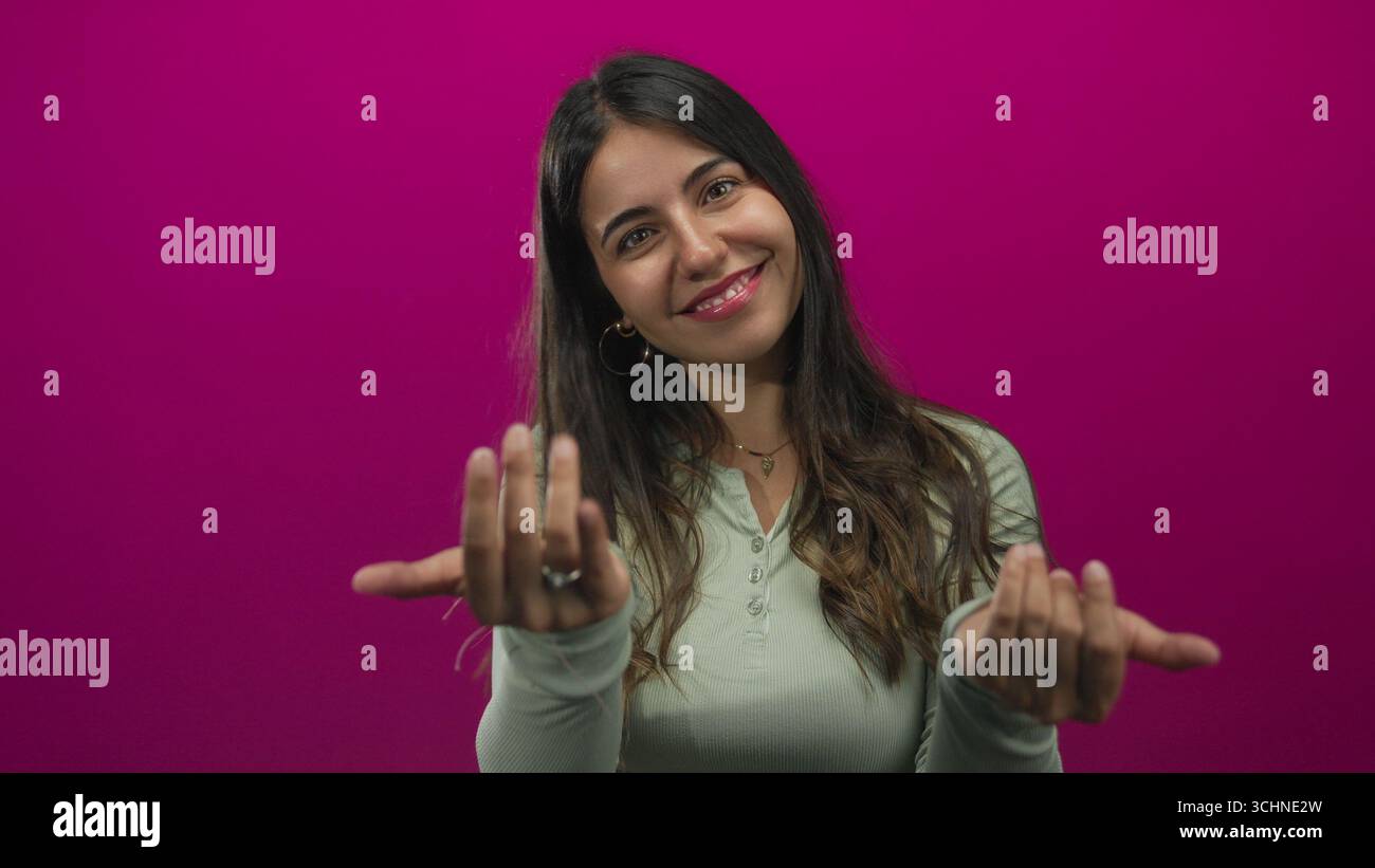 Hispanic woman beckons with open hands in a pink studio with a bright friendly smile; warm invitation. Stock Photo