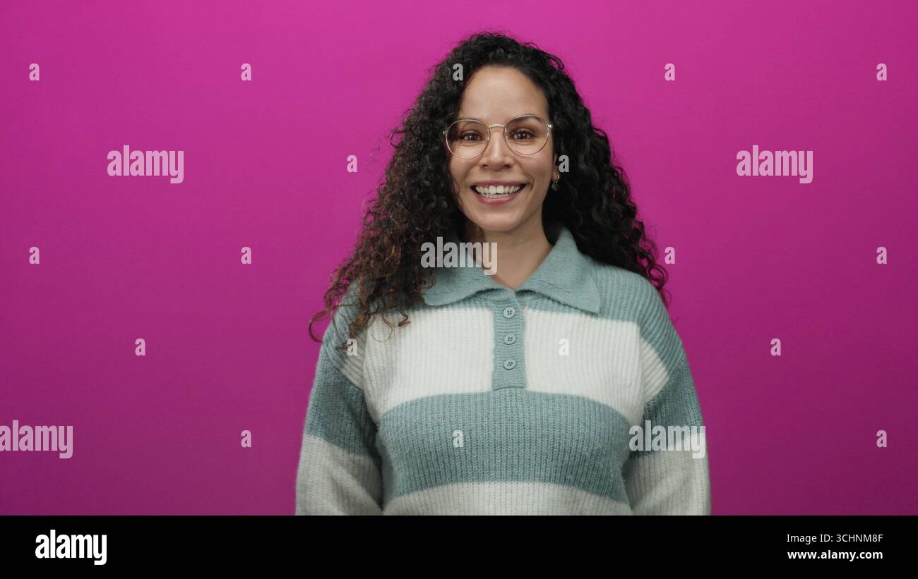 Woman smiling confidently in glasses against vibrant pink background expressing varied emotions with curly hair and warm sweater showcasing casual sty Stock Photo