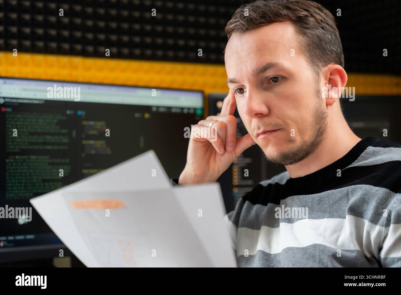 Focused man reading printed documents in software development workspace with code on monitors, reflecting analytical work in tech environment Stock Photo
