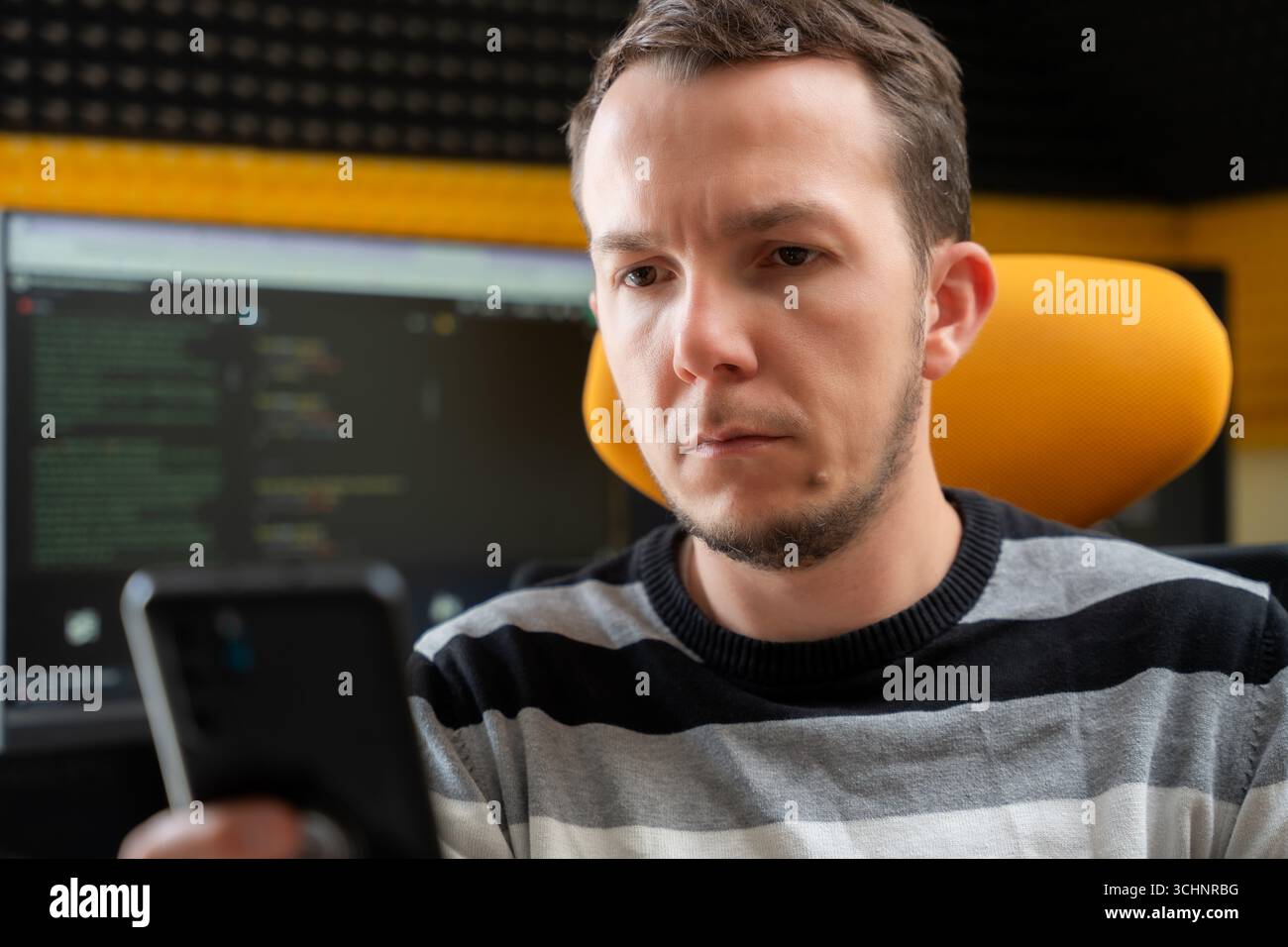 Man looking at smartphone with serious expression in programming workspace, reflecting concentration or tech-related troubleshooting Stock Photo