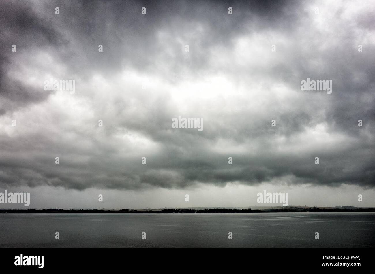 Stormy Clouds Over The Sea Rydebäck Sweden // RYDEBÄCK, Sweden — Stormy, dark clouds hang low over the Öresund strait, with a silhouetted coastline visible in the distance. Rydebäck is a locality situated in Helsingborg Municipality, Skåne County, Sweden. The Öresund is a strait that forms the Danish-Swedish border and is the strait that separates Denmark and Sweden. Stock Photo