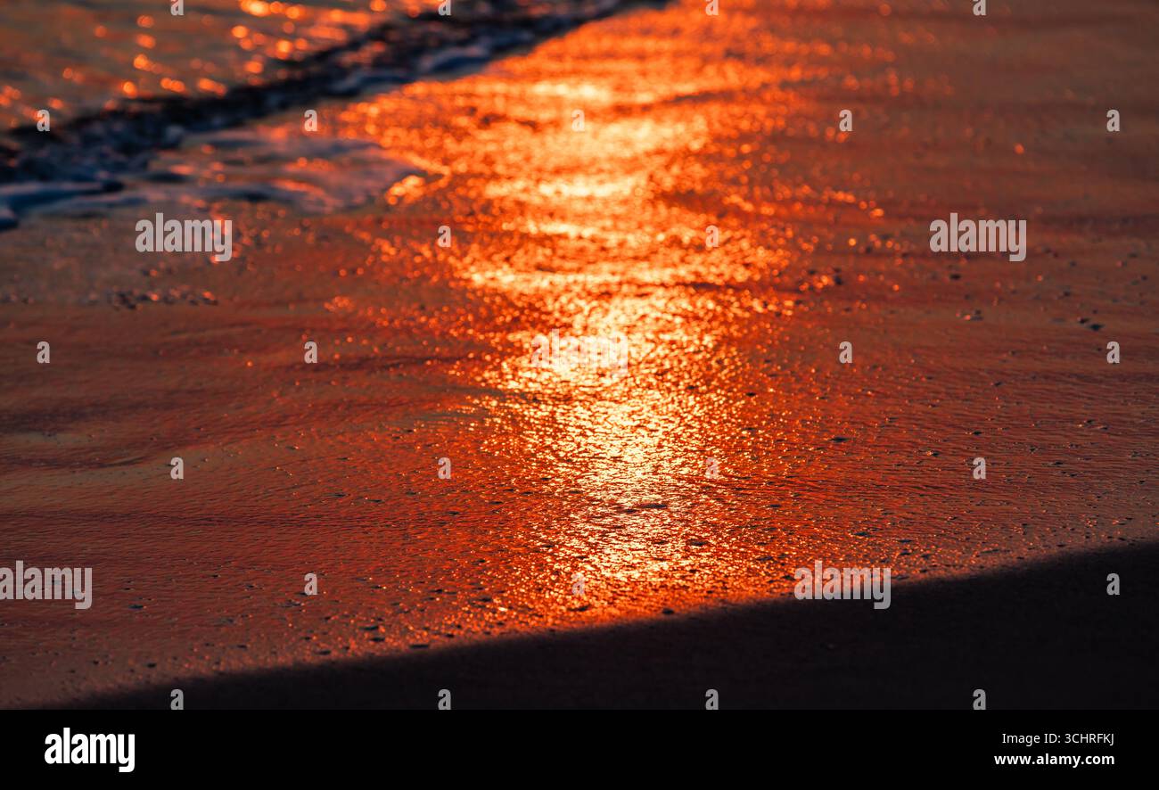 A serene sunset reflecting brilliantly on a sandy shore during low tide, showcasing vibrant orange and red hues along with gentle water ripples, captu Stock Photo