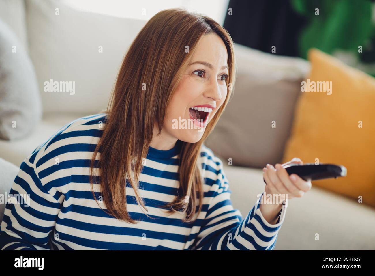 Happy woman in striped sweater enjoying a television show while holding remote in cozy home setting Stock Photo