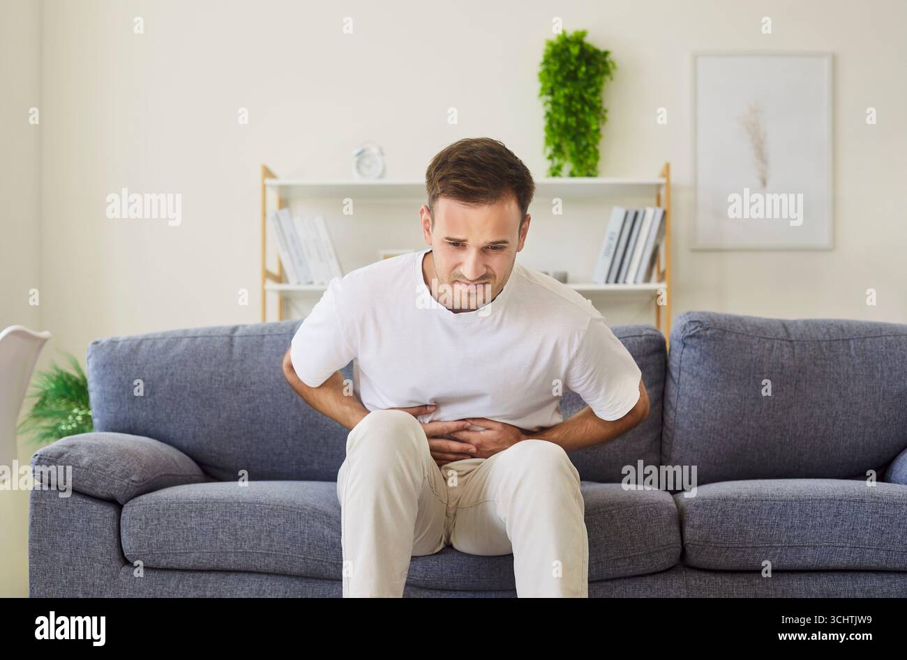 Young man hunched on couch with abdominal pain, pressing stomach in discomfort Stock Photo