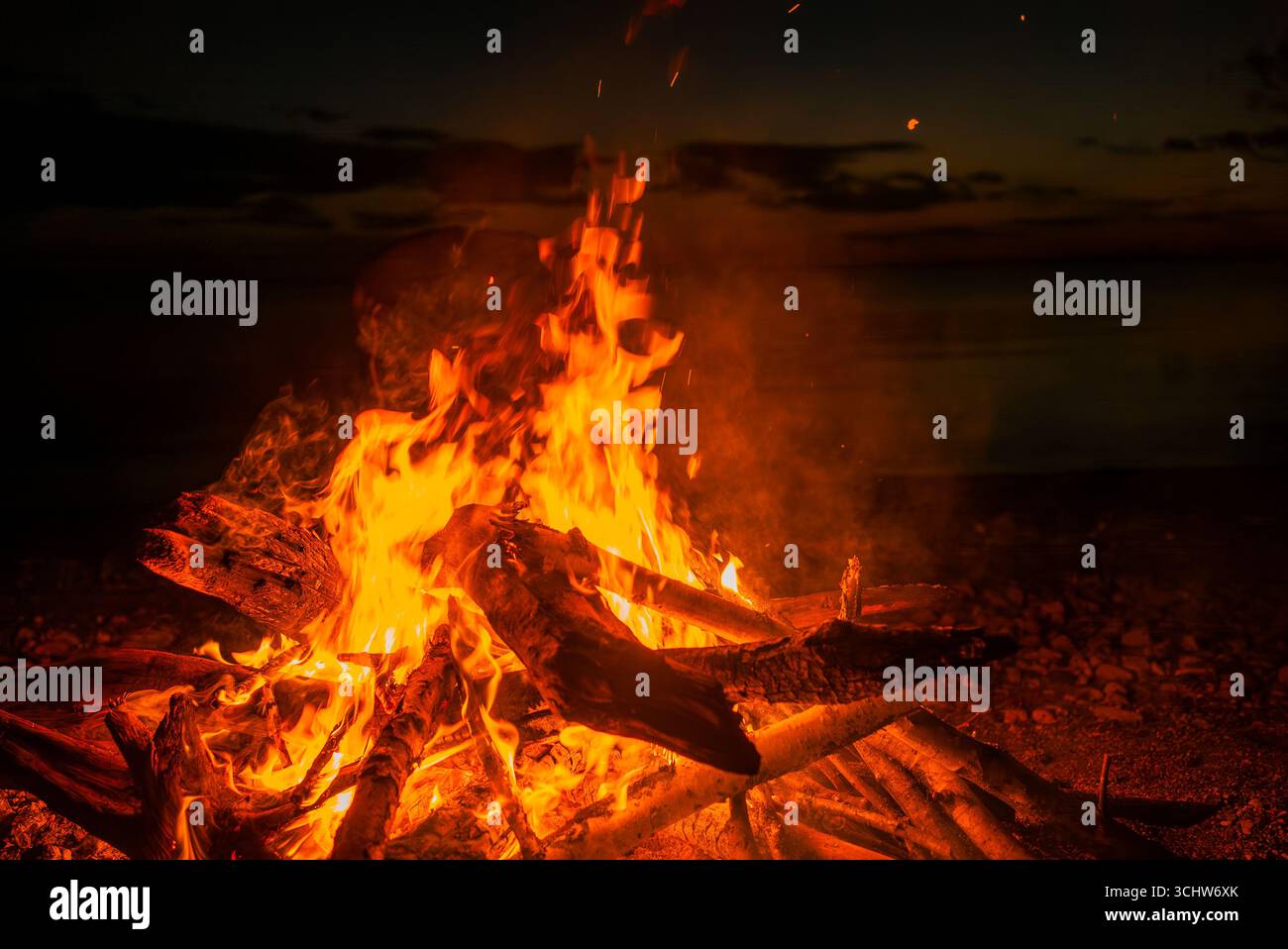 Bonfire burning brightly on a beach at sunset, casting a warm glow across the tranquil waters and creating a captivating spectacle against the darkening sky and distant horizon Stock Photo