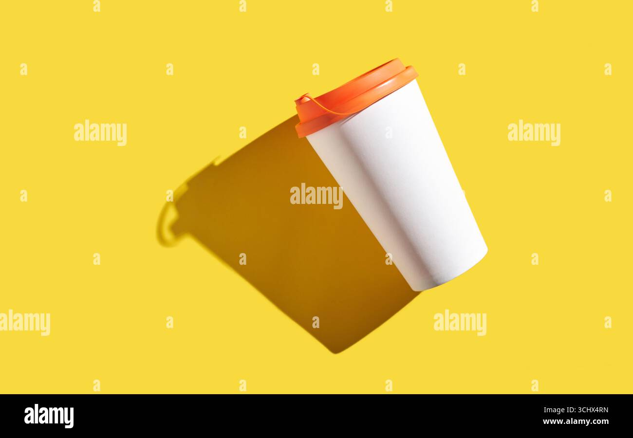 Floating white coffee cup with orange lid on yellow background casting shadow. Stock Photo