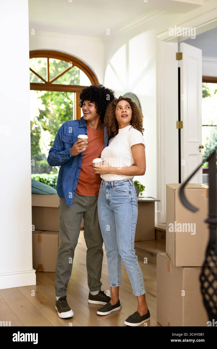 Young diverse couple holding coffee cups, standing among moving boxes at new home Stock Photo