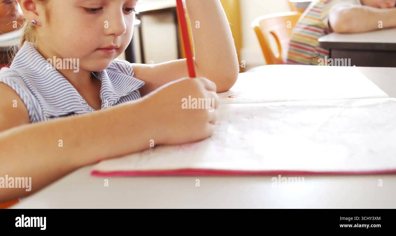 Leaning school-age girl writing in open notebook at classroom desk, with pencil, chair, copy space Stock Photo