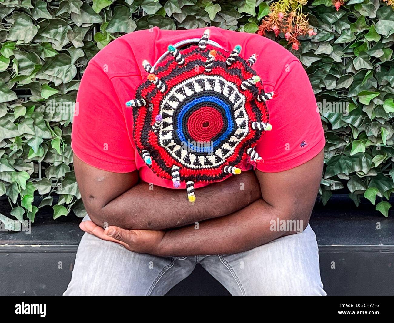 15.06.2023, Berlin, Germany, Europe - A dark-skinned man wearing a colorful knitted hat and crossed arms sits in front of a hedge in Charlottenburg. Stock Photo