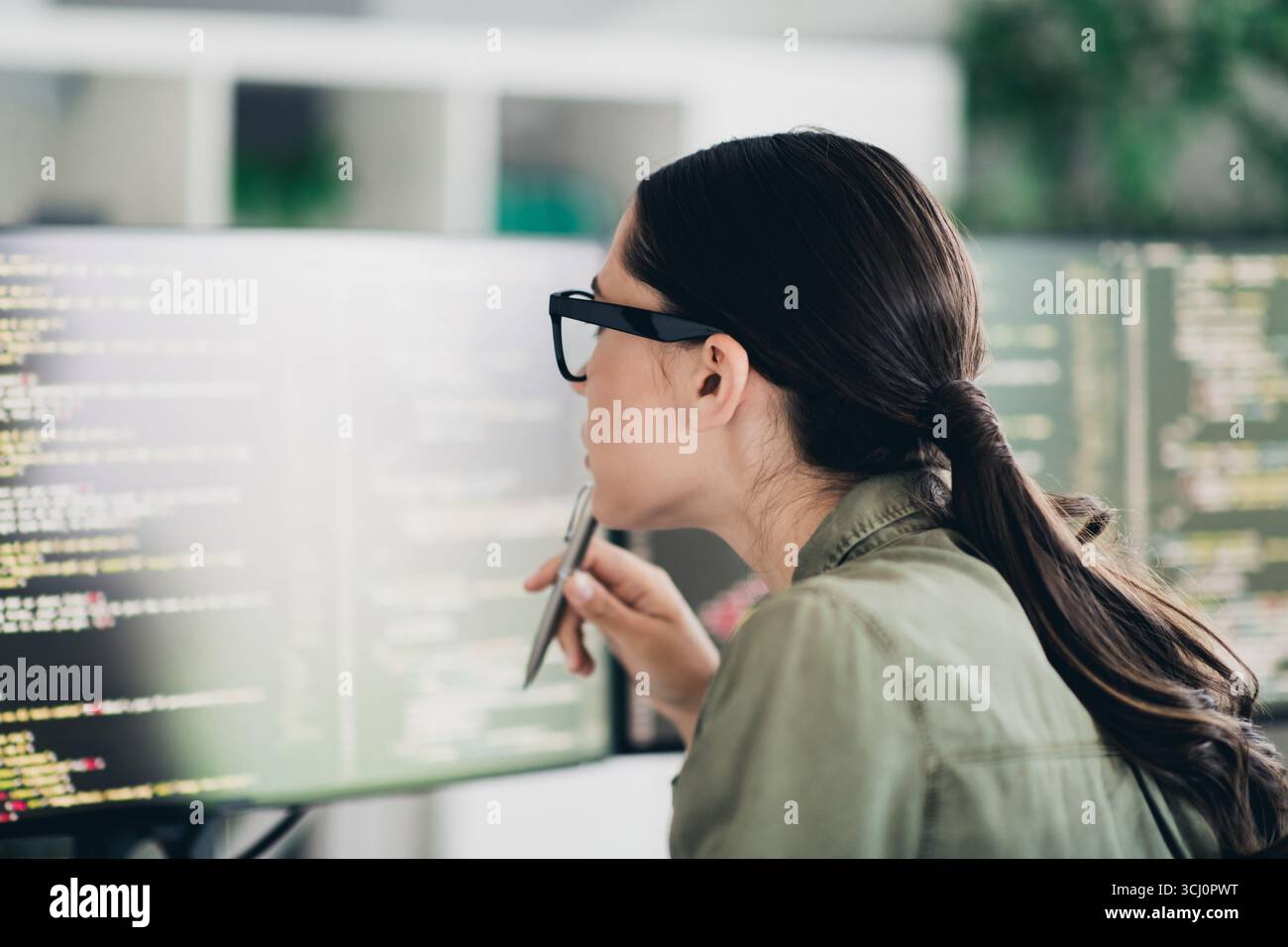 Charming female programmer analyzing code on screen in a modern workspace, focused on debugging and enhancing software Stock Photo