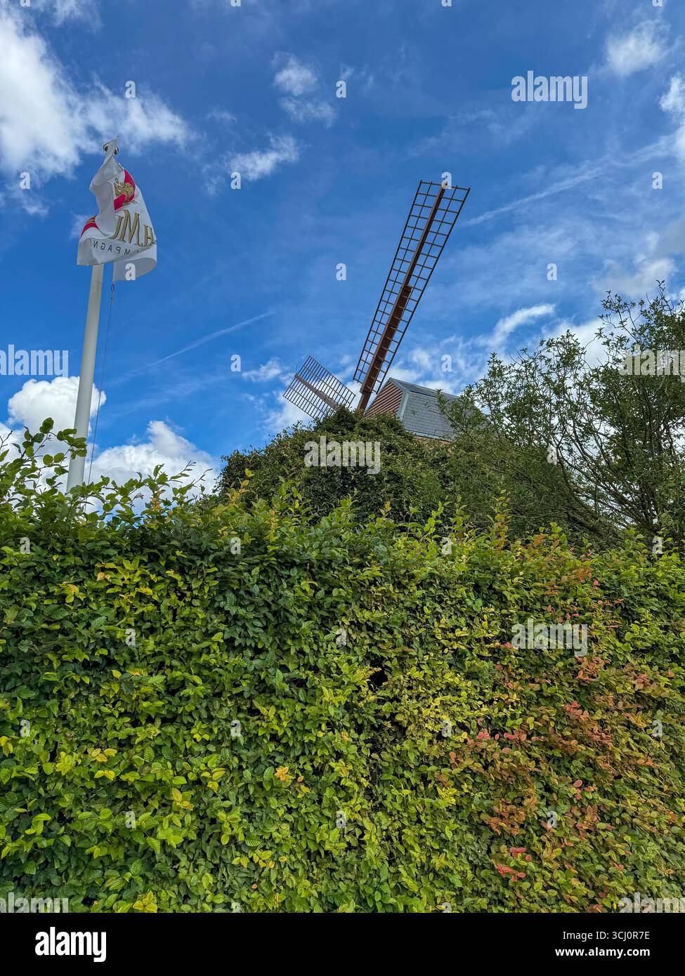 The Verzenay windmill partly hidden by greenery, with th Mumm champagne flag waving in the wind,under a blue sky with clouds, Champagne region, France - Smartphone Captured Stock Image