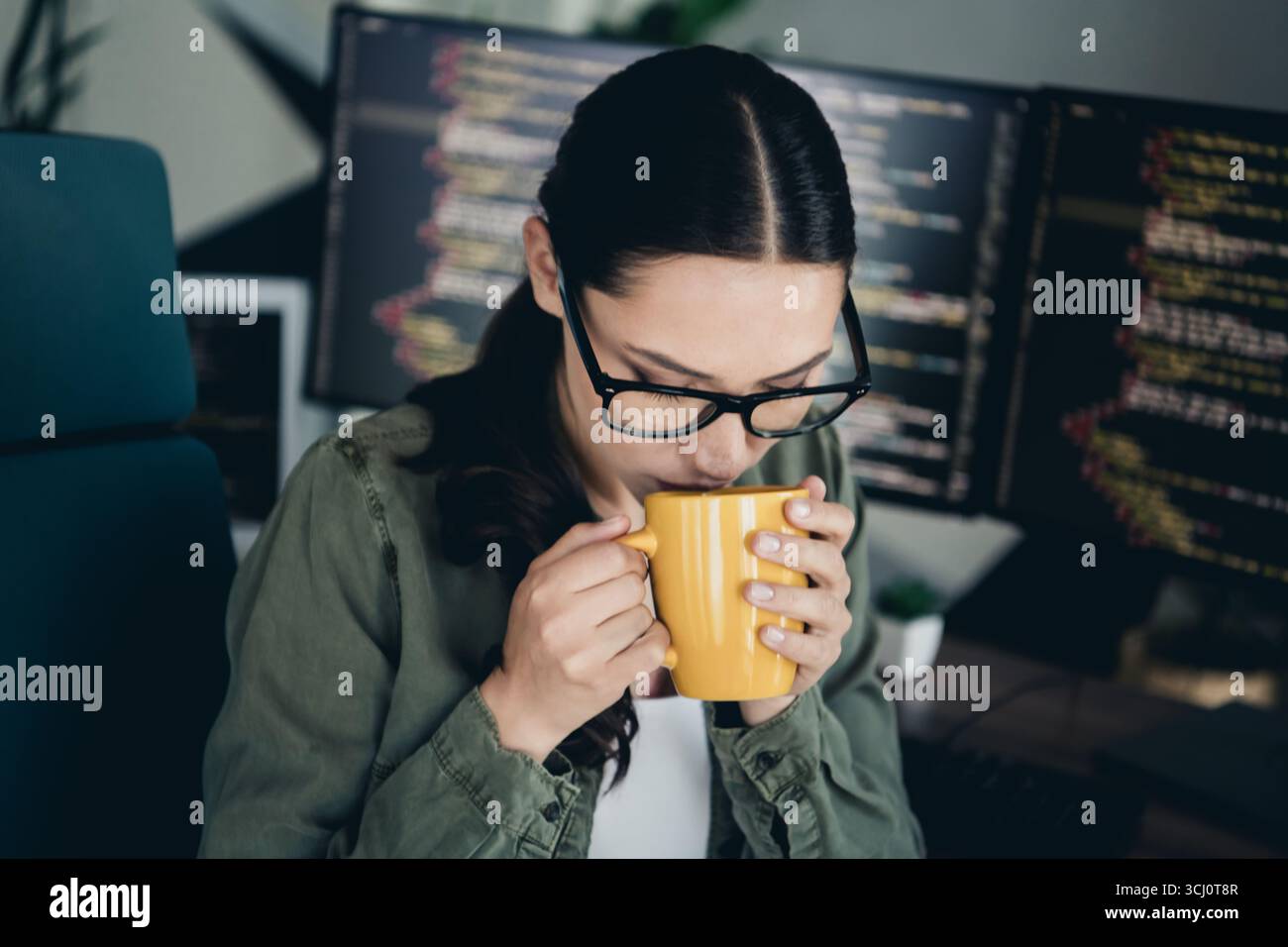 Young female programmer working remotely while enjoying coffee, with computer codes visible on multiple monitors Stock Photo