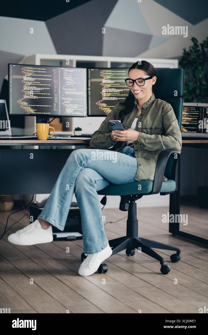 Young female programmer working on a project while using a smartphone in a modern office workspace Stock Photo