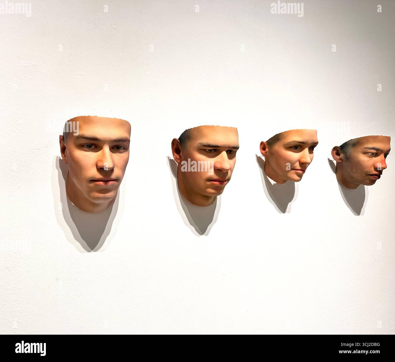 four realistic 3D printed faces mounted on a white wall, creating a striking visual art display with shadows. - Smartphone Captured Stock Image