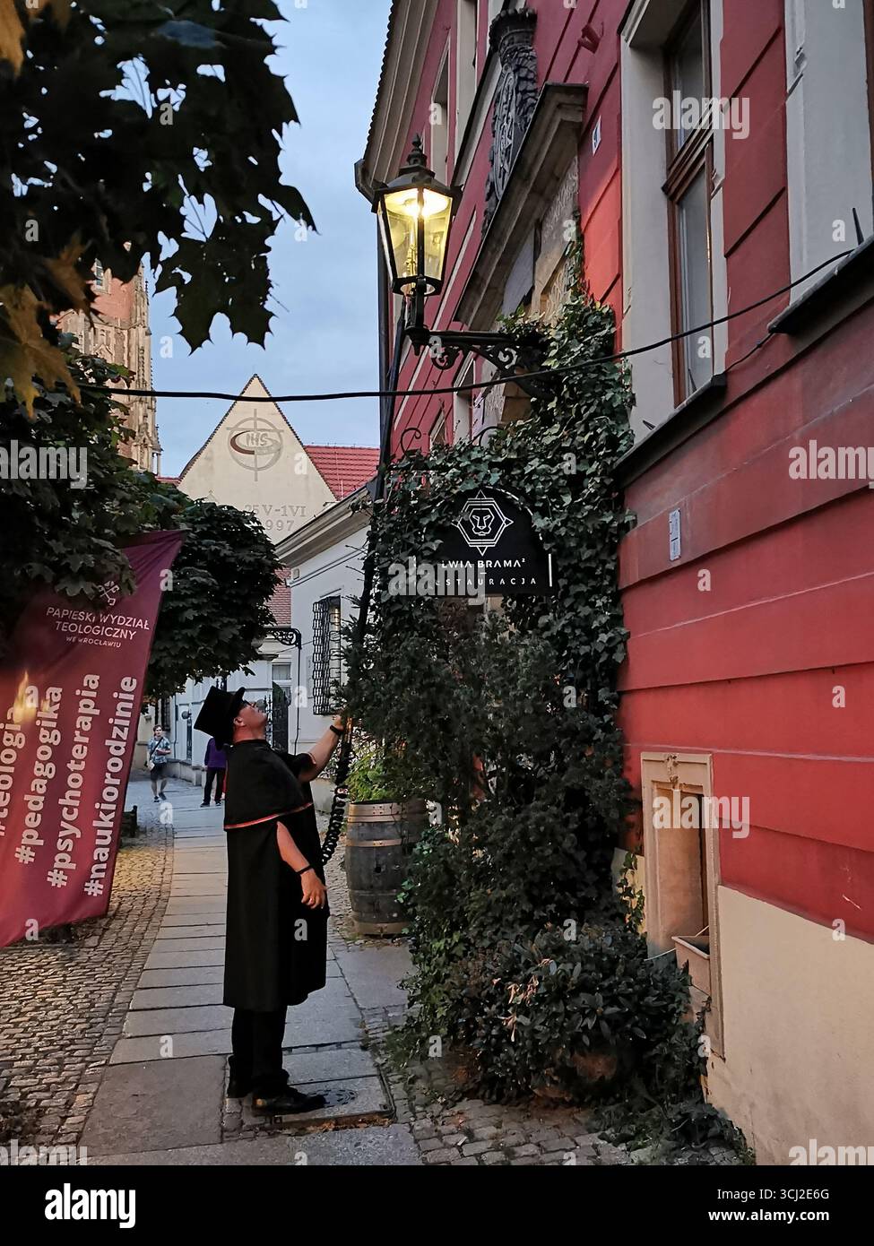 Wrocław lamplighter in hat and cloak lighting gas lamps on Cathedral Island at dusk—historic tradition, Poland, tourism, street scene, Ostrów Tumski. - Smartphone Captured Stock Image