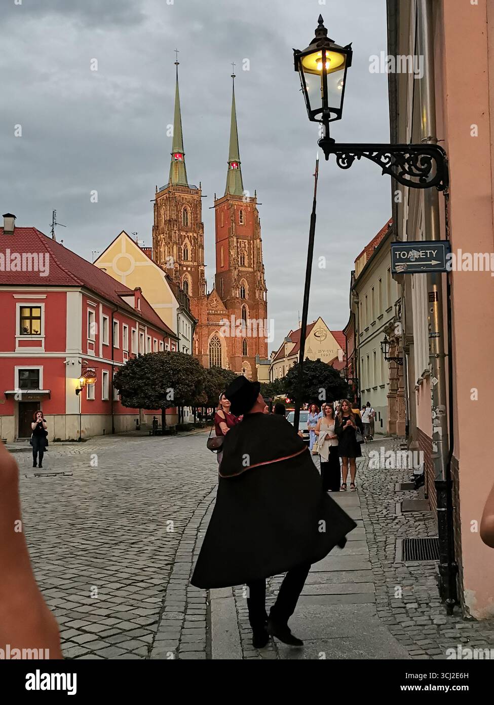 Wrocław lamplighter in hat and cloak lighting gas lamps on Cathedral Island at dusk—historic tradition, Poland, tourism, street scene, Ostrów Tumski. - Smartphone Captured Stock Image