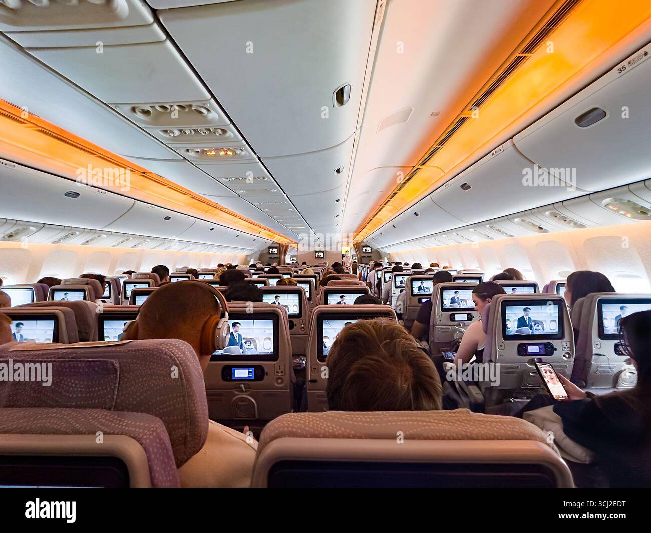 emirates Boeing 777 Airplane cabin filled with passengers watching screens on the back of seats during a daytime flight with orange ambient lighting - Smartphone Captured Stock Image