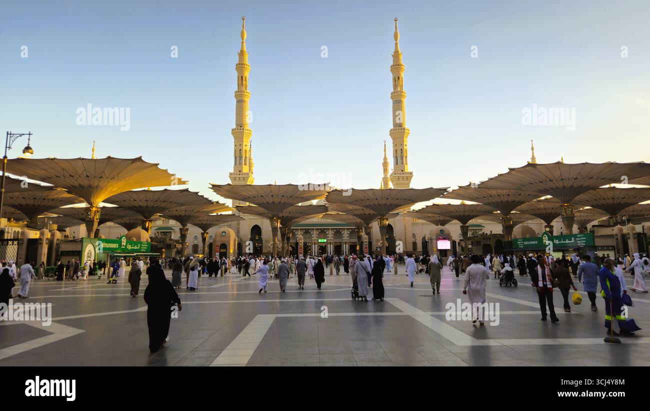 Courtyard of Al Masjid an Nabawi in Medina with minarets, worshippers, and giant umbrellas at sunset. Madinah, Saudi Arabia - March 5th, 2024 - Smartphone Captured Stock Image
