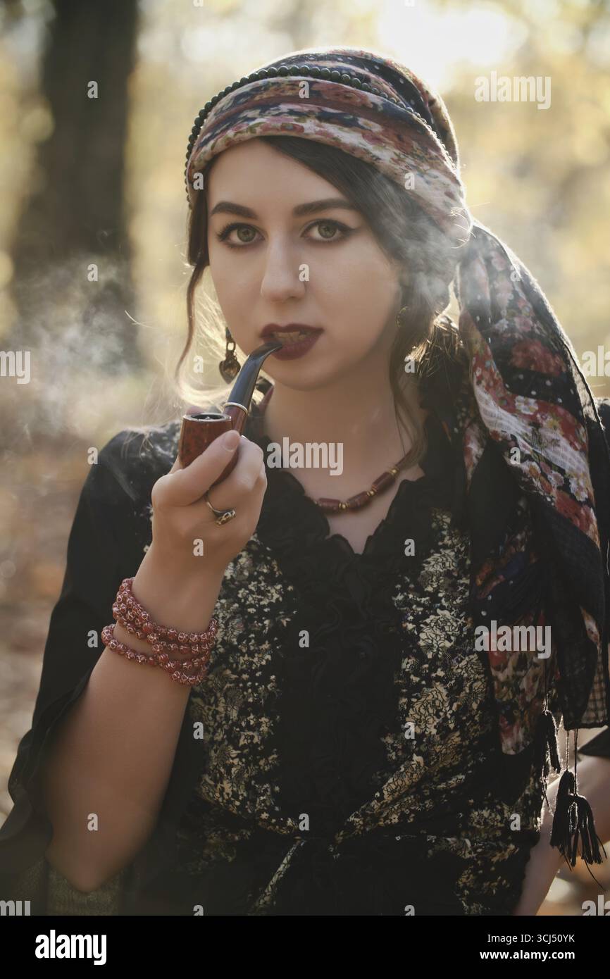 young gypsy woman smoking pipe in autumn forest, looking at camera