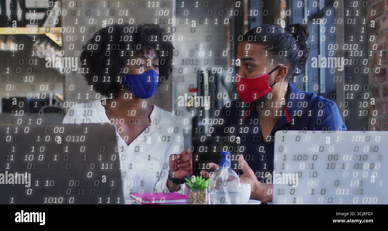 Discussing project two masked colleagues using laptops in cafe, with succulent and binary overlay Stock Photo