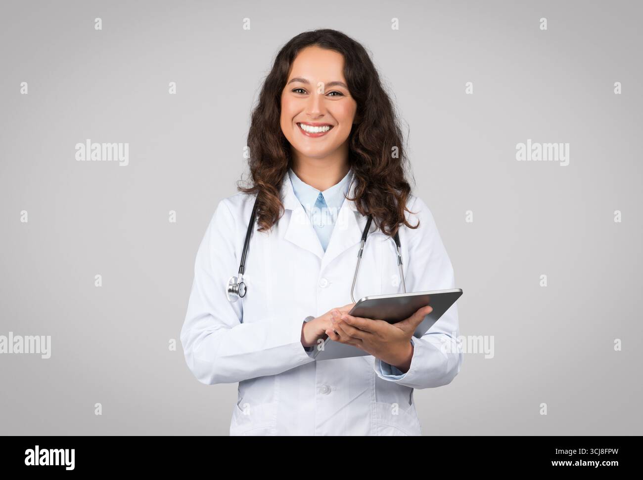 Positive woman in medical workwear doctor using digital tablet isolated on grey background, chatting with patient Stock Photo
