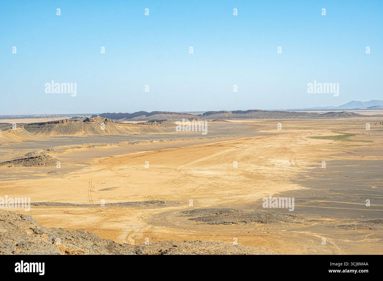 Vast Sahara desert plain in Morocco with golden sand patches dark rocky hills and distant horizon under clear blue sky creating wide panoramic view Stock Photo