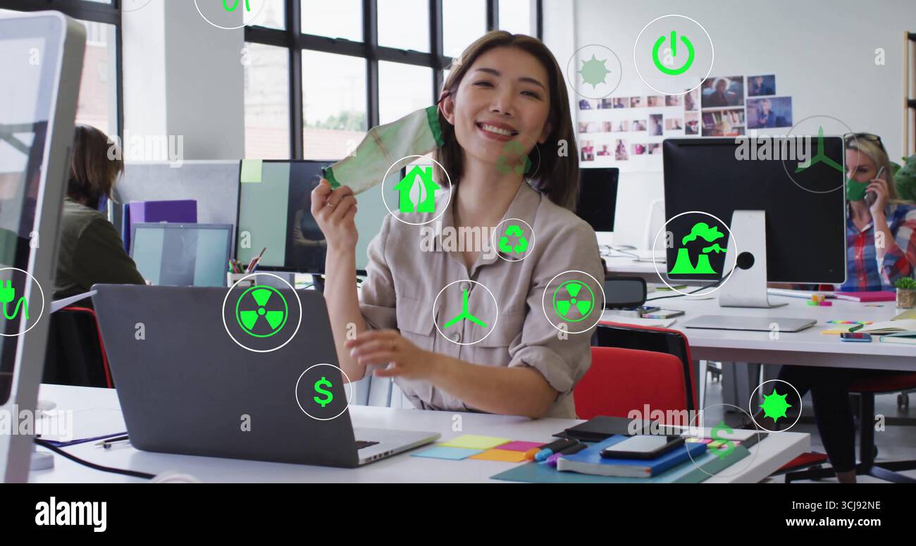 Smiling Asian graphic designer holding green concept card at open-plan office desk, with monitors Stock Photo