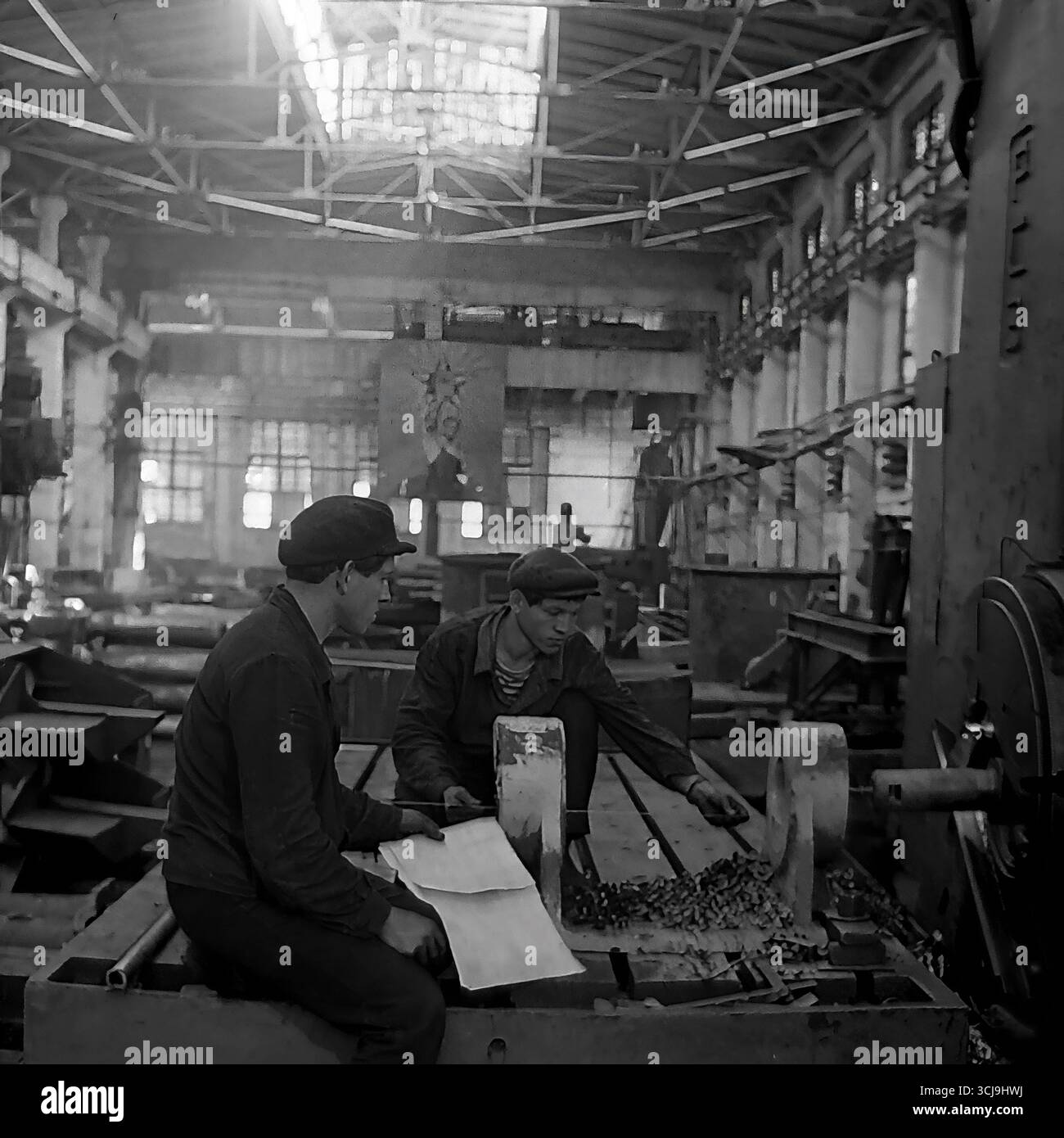 Two Young Workers Study A Blueprint By A Huge Machine Tool In A Heavy Two Young Workers Study A Blueprint By A Huge Machine Tool In A Heavy