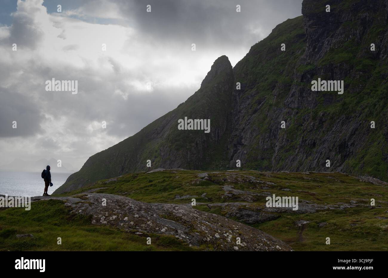 A solitary hiker navigates rocky terrain beside steep cliffs that rise dramatically from the ocean. A village lofoten islands, Norway Stock Photo