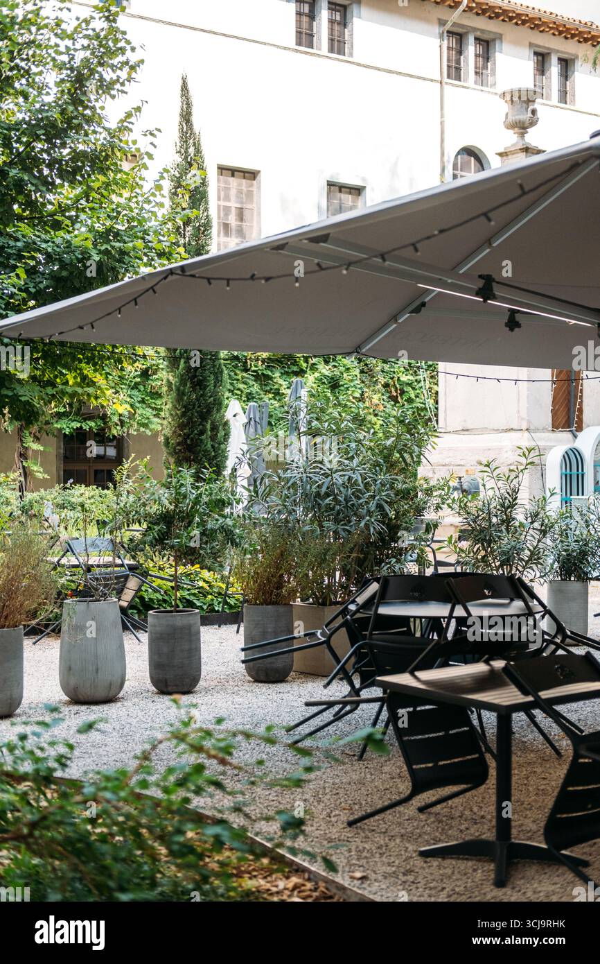 Outdoor garden cafe with large shade umbrella, potted plants, and black chairs in a lush courtyard. Sustainability, urban greenery, social spaces Stock Photo