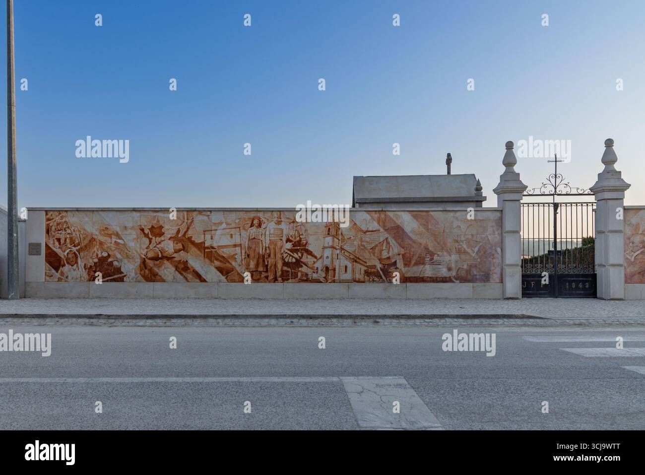 A wide shot of a historical mural in Fátima, Portugal, showing various scenes. The detailed artwork covers a long wall, leading to a decorative gate. Stock Photo