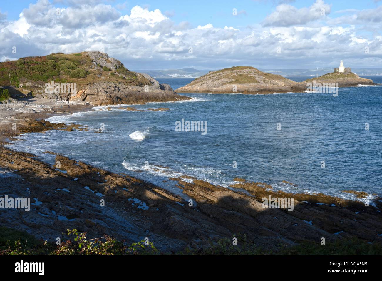 Bracelet Bay, Mumbles - islands at high tide the Lighthouse is situated on the furthest point , waves break on a blue sea reflecting the sunny sky. Stock Photo