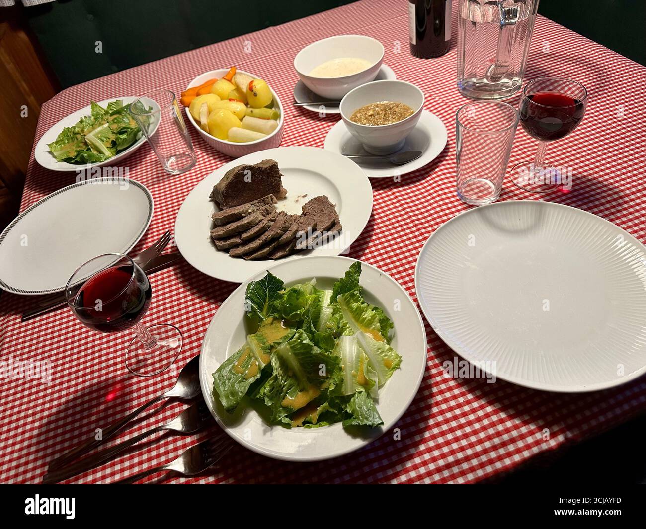 A silverside rump cut or Tafelspitz on a table set for two with sides of green lettuce salad, potatoes, vegetables, two sauces, red wine and water - Smartphone Captured Stock Image