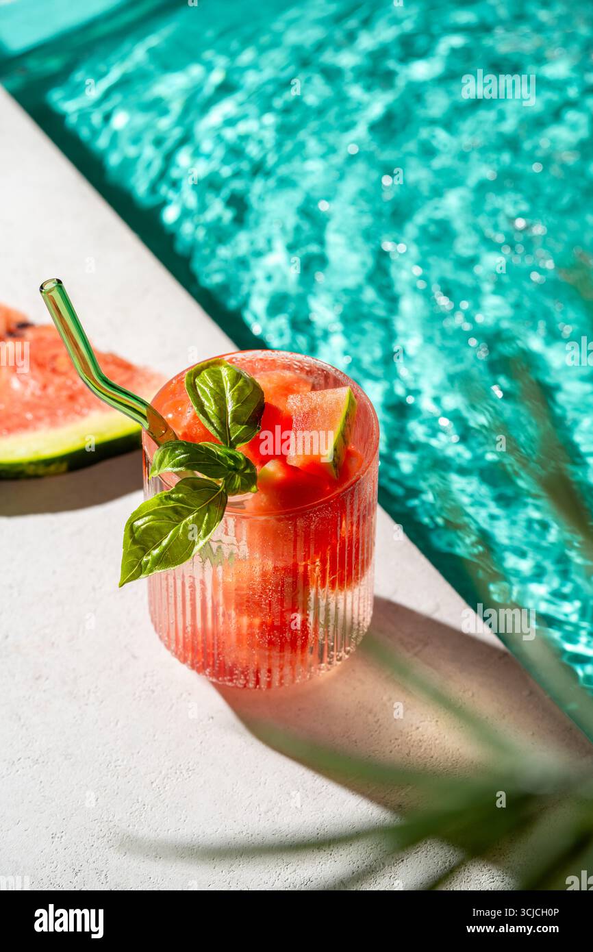 A refreshing watermelon drink in a glass with a green straw, garnished with fresh mint leaves, set on a poolside table. Sunlight reflects off the turq Stock Photo