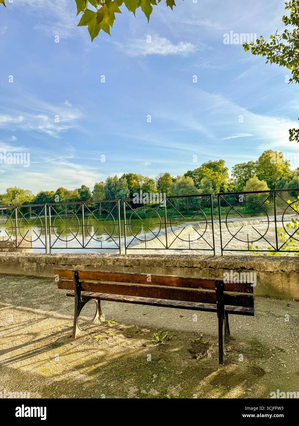 Wooden bench overlooking the Barrage des Forges in Audincourt, France ,with decorative railing and reflections of green trees on the calm water - Smartphone Captured Stock Image