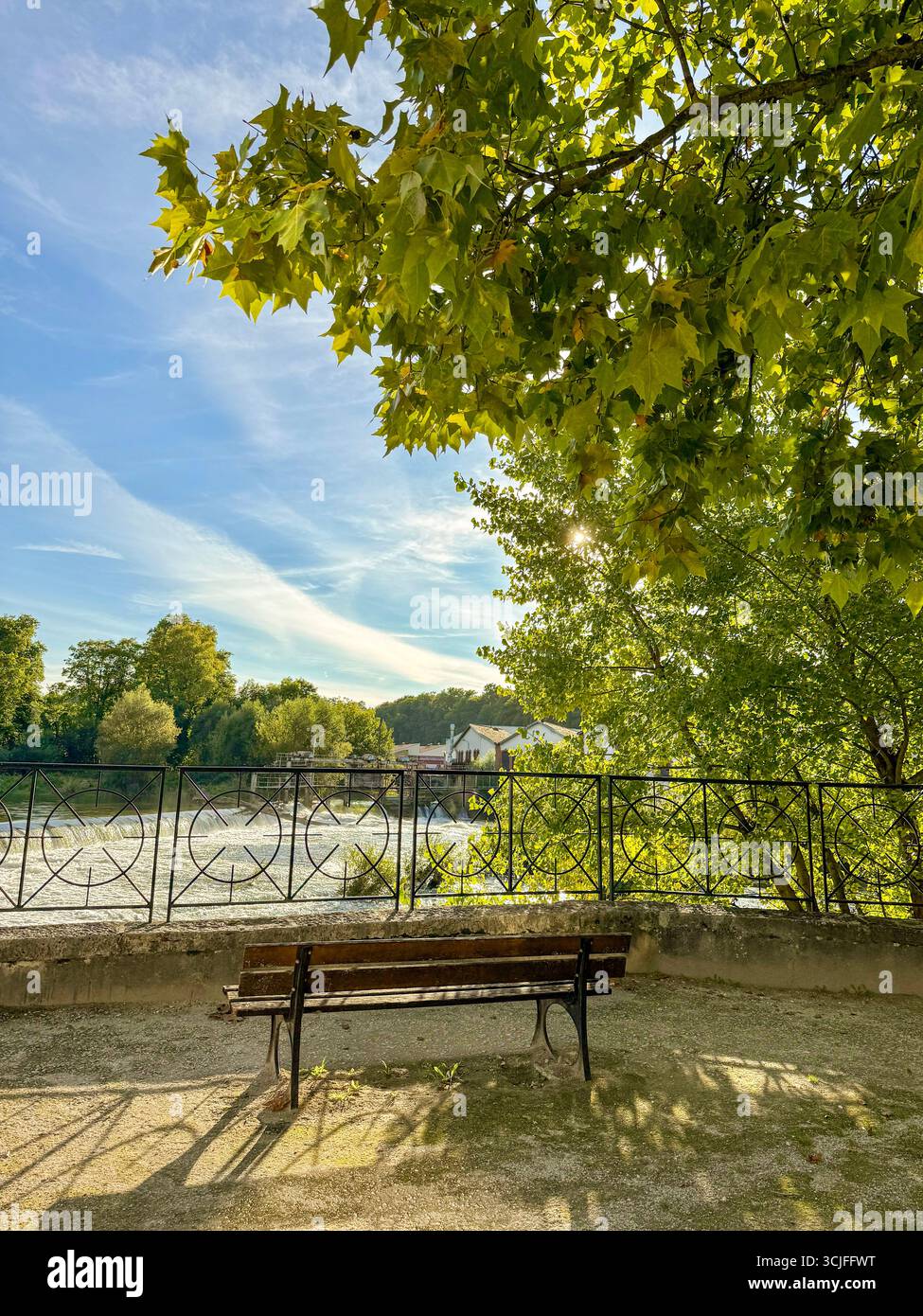 Wooden bench overlooking the barrage des Forges, with metal railing, trees and an old building in the background, under soft sunlight - Smartphone Captured Stock Image
